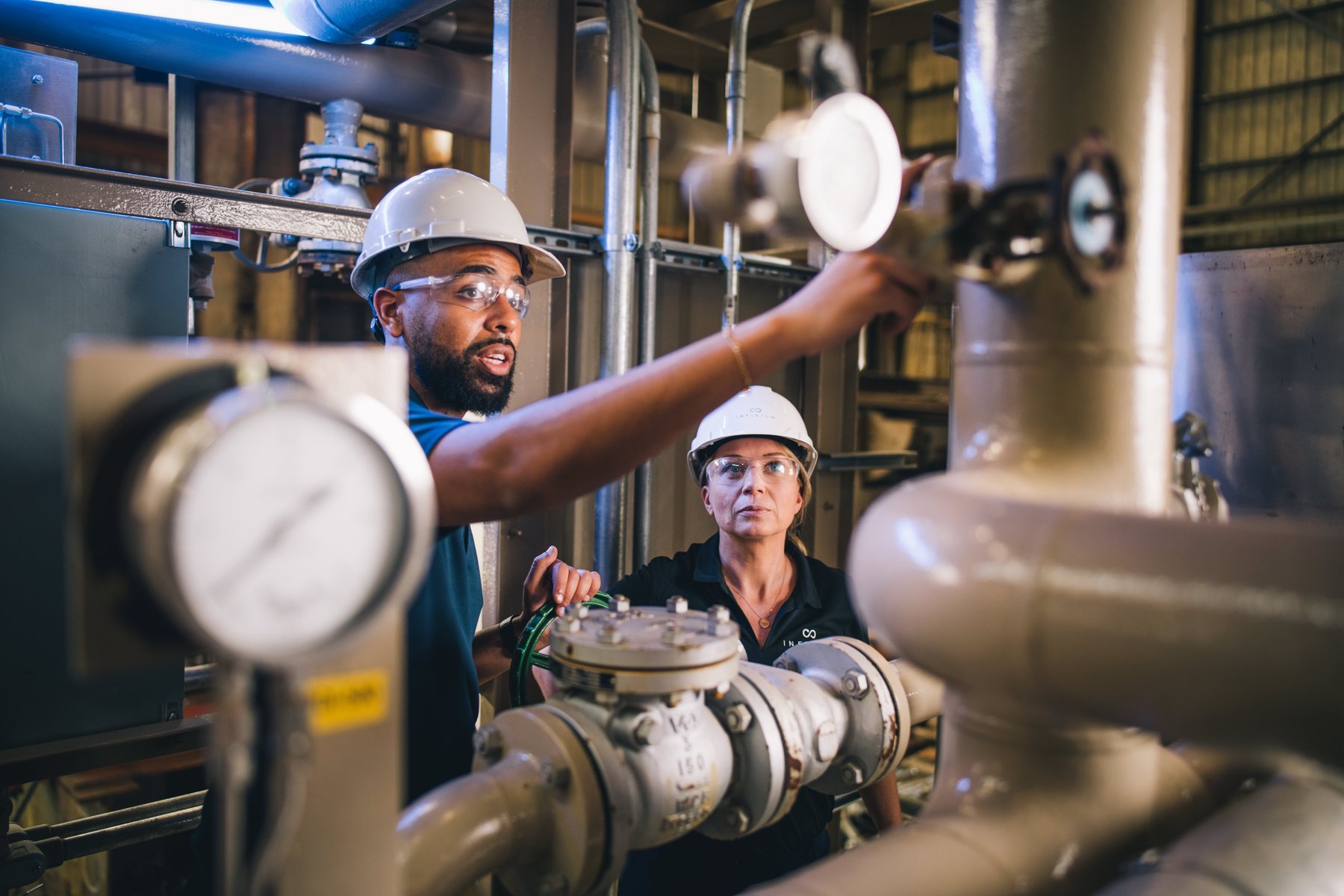 Two Infinium engineers in hard hats and safety glasses checking valves and a pressure gauge on a process skid